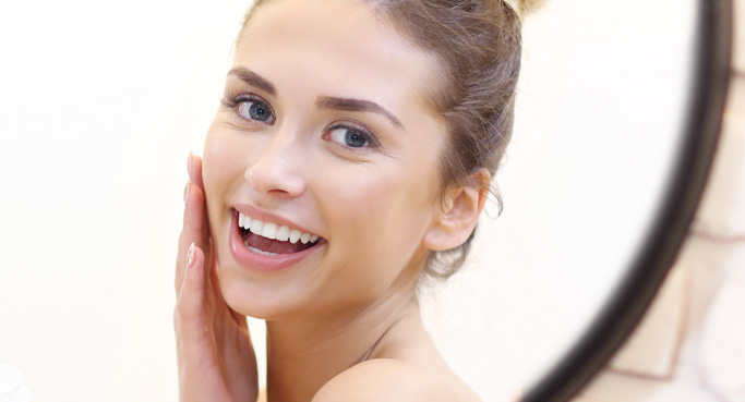 Lächelnde Frau pflegt ihr Gesicht vor dem Spiegel. Steht für Regeneration, Schutz und intensive Hautpflege mit NEOVITA Produkten.